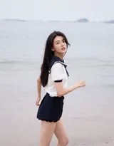 梁洁侧身坐在湿润沙滩上，低头浅笑，黑色短裤衬托出修长腿部线条，脚边清澈海浪轻轻涌来又退去